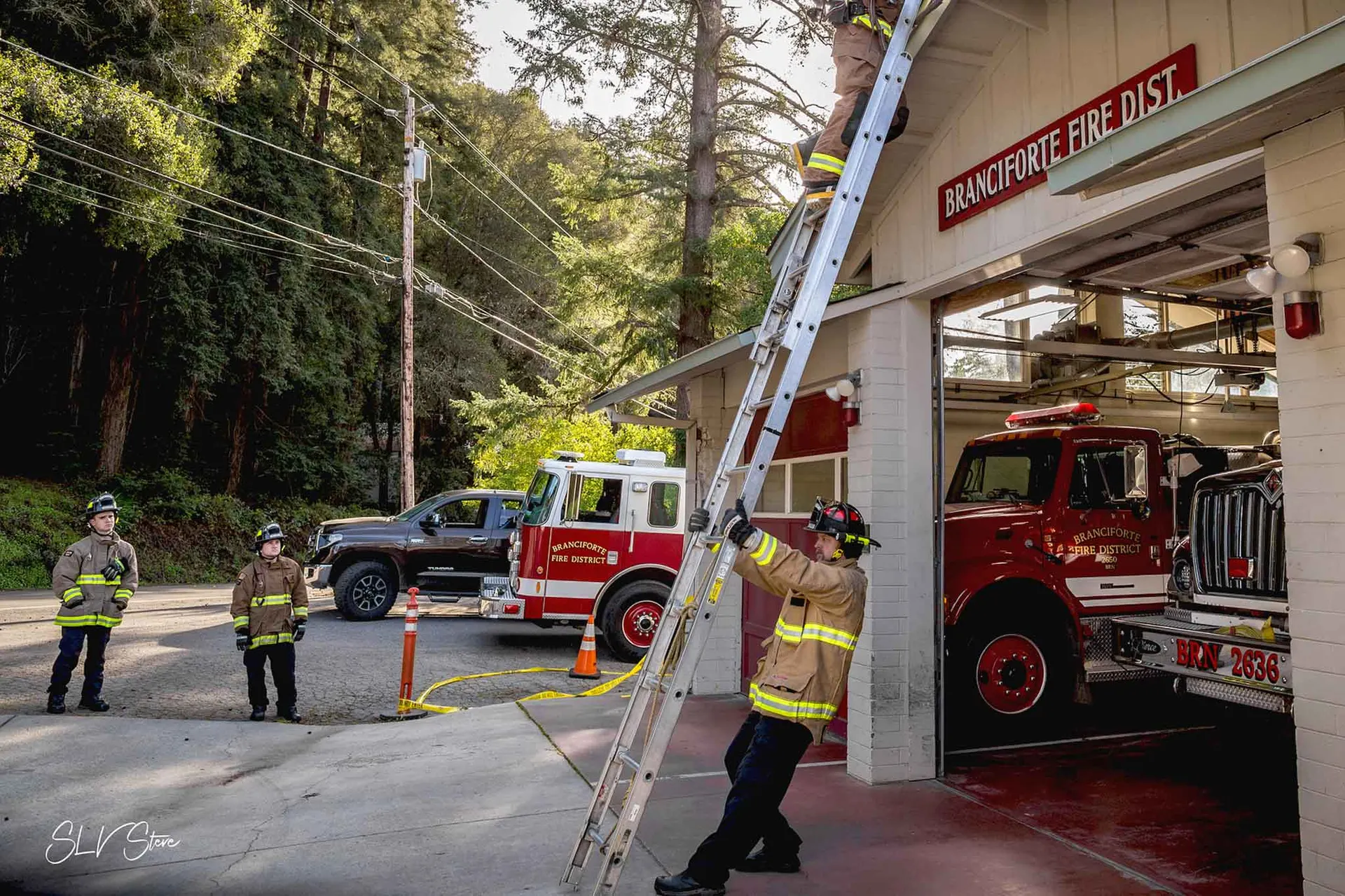 Two firefighters observing firefighter holding ladder while another firefighter climbs to roof of Branciforte Fire Station.